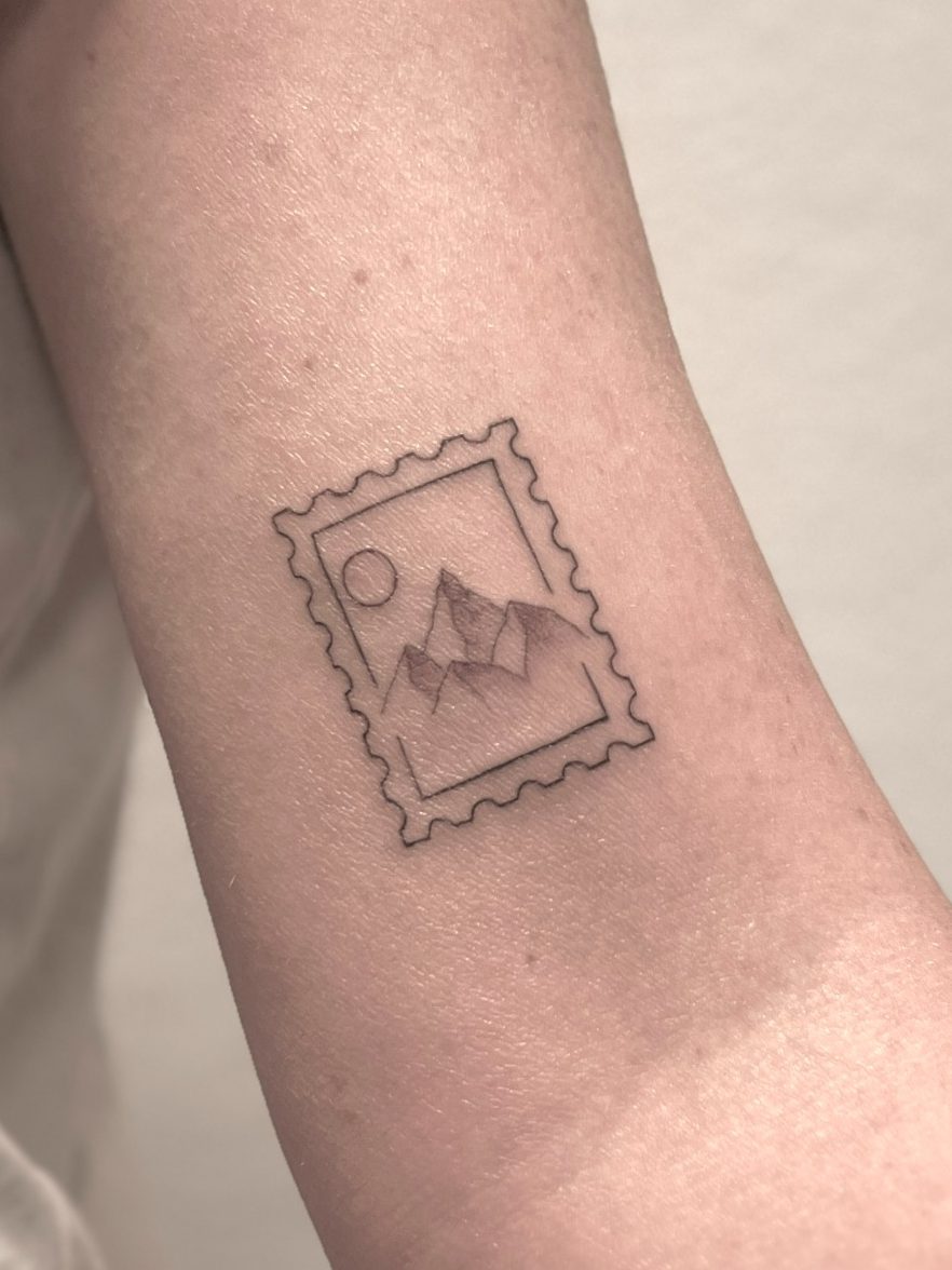 Tattoo eines Briefmarkenmotivs mit einer Berglandschaft und Sonne – aus dem Studio Rabauke Tattoo and Arts in Neu-Ulm