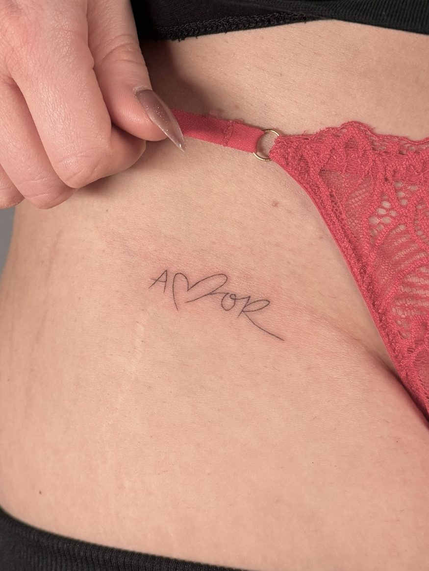 Tattoo mit filigraner Schrift "Amor" auf der Hüfte, sichtbar unter roter Unterwäsche – tätowiert in Neu-Ulm | Ink Shell