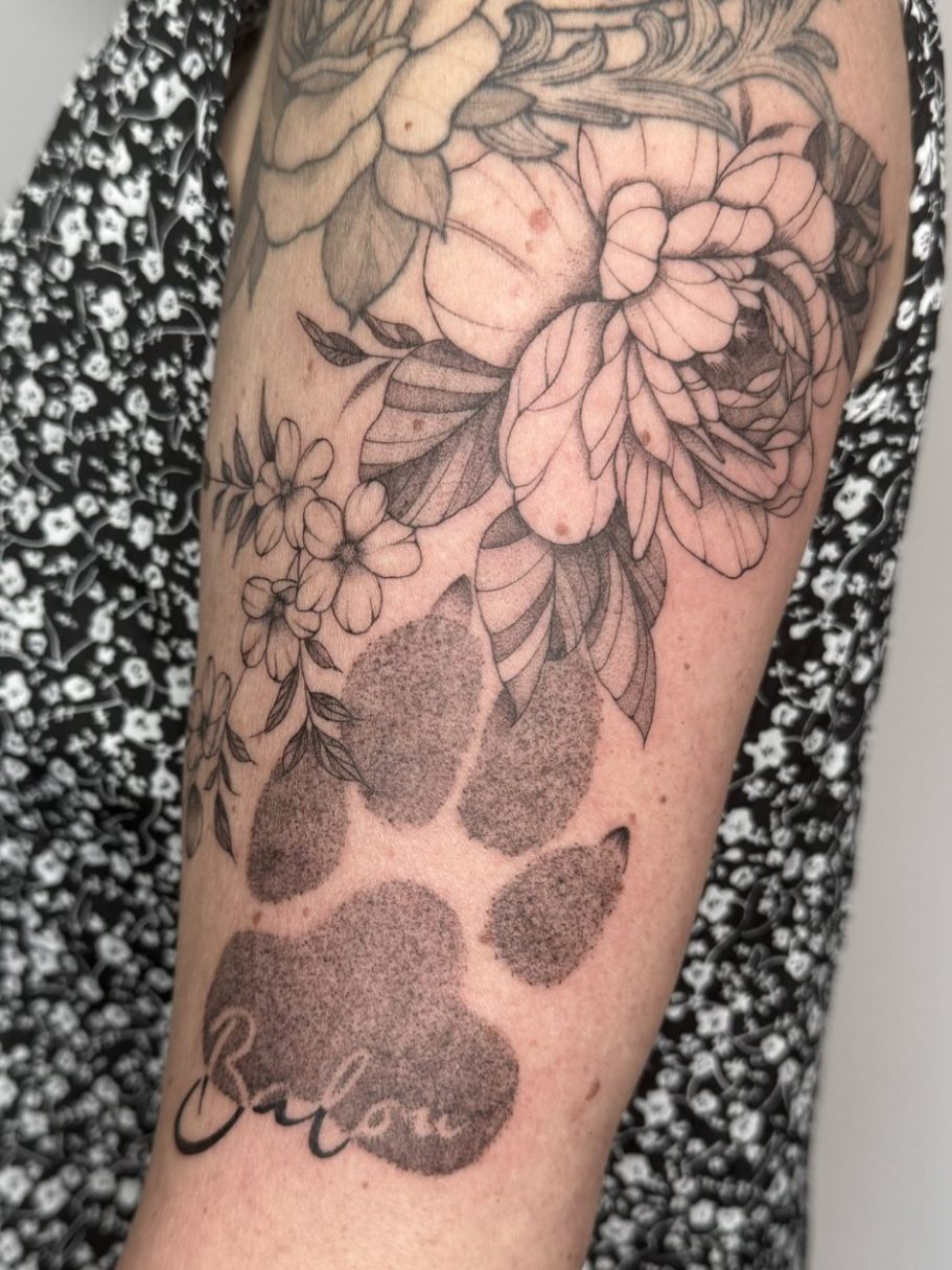 Tattoo einer Dotwork Hundepfote und Fineline Blüten mit Name des Hundes auf einem Oberarm – Ink Shell Tattoo Studio in Neu-Ulm